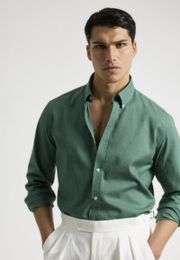 Camisa verde de botones hecha de lino, con mangas largas arremangadas, combinada con pantalones blancos de cintura alta y un tatuaje visible en el pecho.