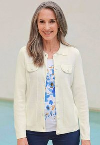 Lichtcrème cardigan met knoopsluiting aan de voorkant, twee borstzakken en een geribbelde textuur, gelaagd over een witte blouse met bloemenpatroon en donkere broek.