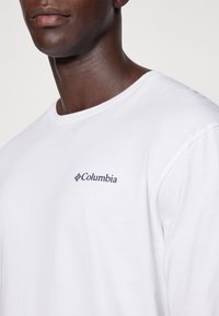 Valge pika varrukaga T-särk, mis on valmistatud puuvillast. Vasakul rinnal on sinine Columbia logo. Lihtne ümara kaelusega disain ja tavaline lõige.