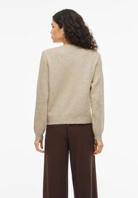 Šviesiai smėlio spalvos megztinis su apvalia iškirpte ir ilgomis rankovėmis, pagamintas iš minkštos medžiagos. Turi kūgines apačias ir rankogalius, dėvimas kartu su rudomis kelnėmis.