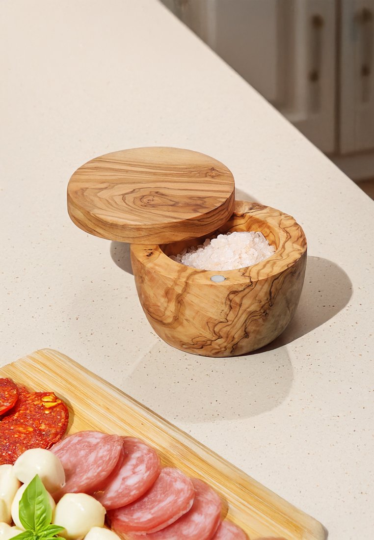 Drewniana solniczka o okrągłym kształcie i płaskiej pokrywce. Drewno oliwne ma naturalny wzór słojów i zawiera gruboziarnistą sól w środku.