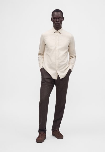 Homme debout les mains dans les poches, portant une chemise beige à manches longues, un pantalon marron foncé et des chaussures marron assorties sur fond uni.