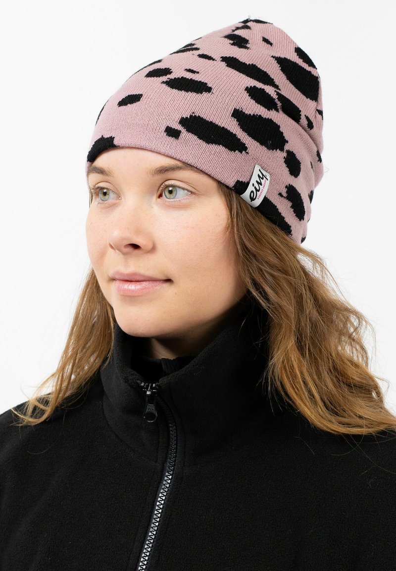Een beanie in lichtroze met zwarte abstracte vlekken. Gemaakt van gebreid materiaal, met een label met logo. Draag het met een zwarte fleece jas.