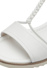 Sandale blanche avec une large bride en matériau synthétique lisse, une bride tressée et une semelle beige texturée.