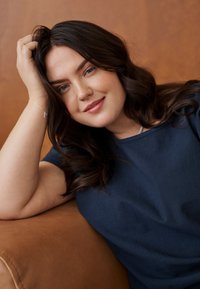 Jonge vrouw met lang donker haar en blauwe ogen leunend op een bruin bank, met haar hoofd rustend op haar hand, gekleed in een marineblauw shirt.