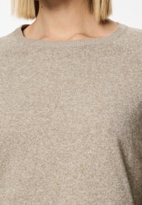 Beige strikket sweater båret af en person med kort blont hår, som viser halsudskæringen og den øverste del af torsoen.