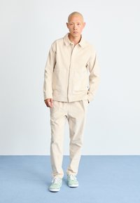 Uomo che indossa una giacca e pantaloni beige con sneakers verdi e bianche, in piedi su un pavimento grigio contro uno sfondo bianco.