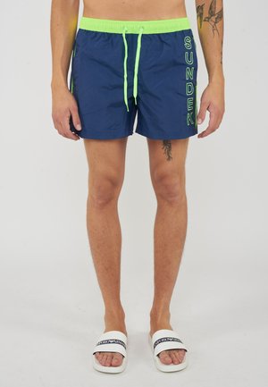 Mann trägt marineblaue Badeshorts mit neongrünem Bund und Kordeln, "SUNDEK" Text am rechten Bein und weißen Emporio Armani Sandalen.