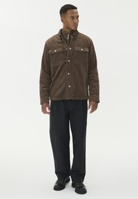 Pruunist korduroist jakk kahe rindkere tasku ja näpitsnuppudega, koos mustade pükstega ja mustade lipsutavade kingadega.