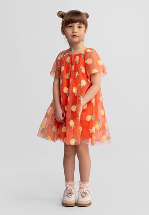 Jeune fille aux cheveux bruns coiffés en deux chignons, portant une robe orange à motifs de fruits jaunes, des manches transparentes, des chaussettes roses à volants et des chaussures blanches.