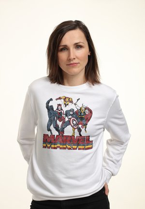 Kobieta z brązowymi włosami do ramion, ubrana w białą bluzę Marvel z postaciami Black Panther, Scarlet Witch, Iron Man, Captain America i Thor.