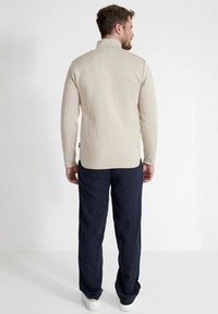 Beige Langarmshirt mit hohem Kragen und gerippter Textur, kombiniert mit dunkelblauen, locker sitzenden Hosen und weißen Sneakers.