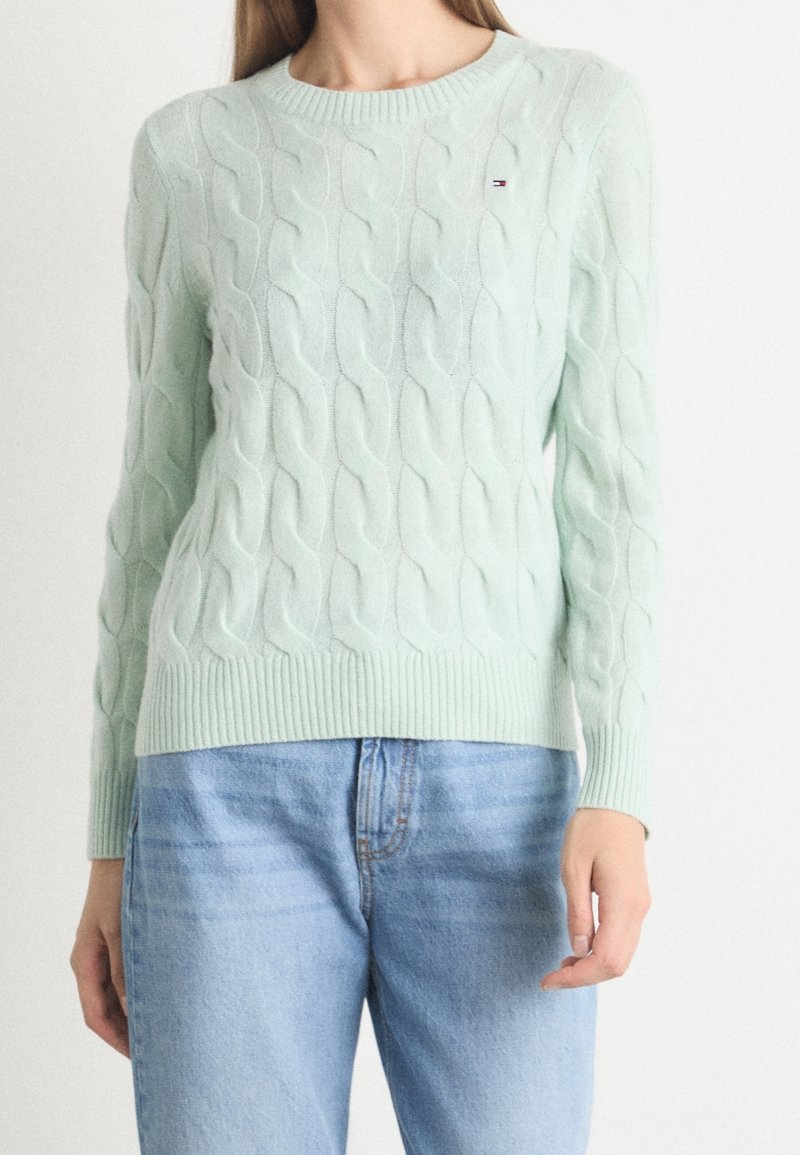 Femme portant un pull torsadé vert clair associé à un jean bleu clair, debout devant un fond blanc uni.