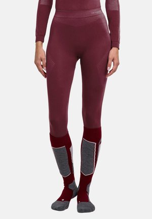Persoon die strakke bordeauxkleurige leggings en bijpassende lange sokken draagt met grijze en witte gewatteerde gedeelten, staand tegen een effen achtergrond.