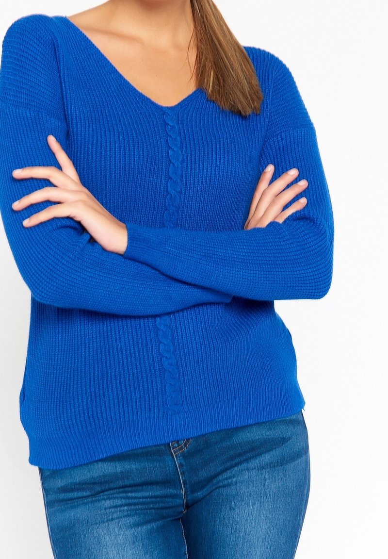 Blå stickad tröja med V-ringning och fläktstickade detaljer längs framsidan. Mjuk textur och långa ärmar, kombinerad med denimjeans.