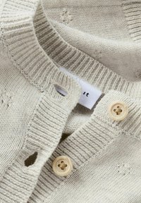 Cardigan a maglia di un leggero beige con colletto a coste, due bottoni di legno e una piccola etichetta con il nome del marchio visibile.