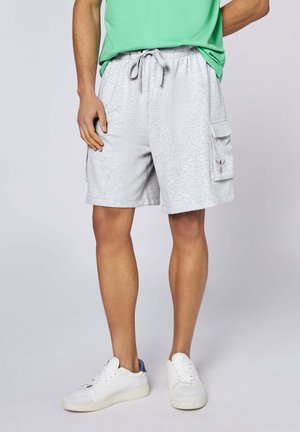 Mann trägt hellgraue Cargo-Shorts mit Seitentaschen, ein grünes Hemd und weiße Sneakers vor einfachem Hintergrund.