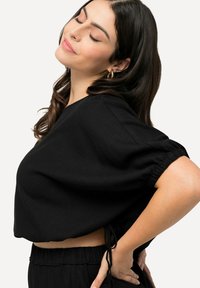Top noir court avec des manches bouffantes courtes, fabriqué dans un tissu texturé. Présente des liens sur les côtés à la taille pour un ajustement réglable.