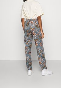 Henkilö, jolla on päällä siniset kukkakuosiset housut, valkoinen lyhythihainen paita, valkoiset sukat ja valkoiset tenniskenkiä, seisoo yksinkertaista taustaa vasten.