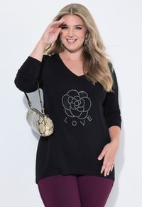 Uśmiechnięta kobieta z długimi blond włosami nosi czarną bluzkę z kamyczkami w kształcie kwiatu i napisem "LOVE", fioletowe spodnie oraz torebkę we wzór wężowej skóry.