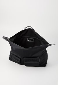Sac en nylon noir avec fermeture éclair, comportant un grand compartiment et deux poches avant. L'intérieur affiche une étiquette de marque. Texture lisse.
