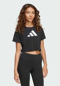 Crna skraćena majica s bijelim Adidas logotipom, kratki rukavi, okrugli izrez, uparena s crnim tajicama visokog struka. Glatka tekstura.