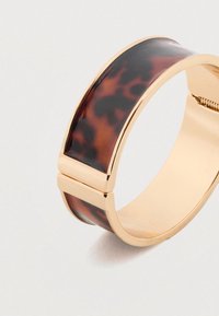 Surašas torto uždaro bangles, išsiskiriantis šukuotės raštu, su auksine metaline akcento ir uždarymo sistema, lygia tekstūra ir apvaliais kraštais.