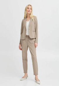 Beige maßgeschneiderter Blazer mit einem Knopf und zwei vorderen Taschen, kombiniert mit passenden schmalen Hosen. Eine weiße Bluse und spitze Ballerinas runden das Outfit ab.