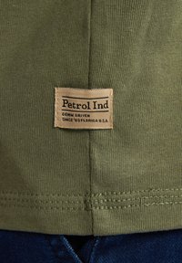 Olive green t-shirt with textured fabric, featuring a tan label stitched on the hem that reads "Petrol Ind, Denim Driven, Since '89 Florida U.S.A."
