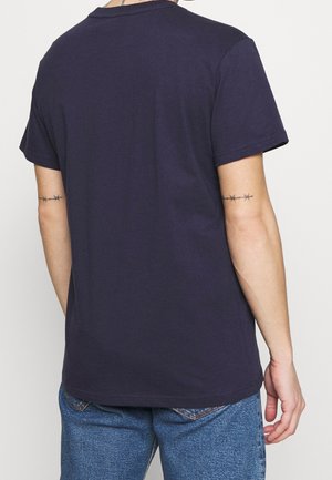 Personne portant un t-shirt bleu marine uni à manches courtes et un jean bleu, vue de dos avec des tatouages de fil barbelé sur les deux bras supérieurs.