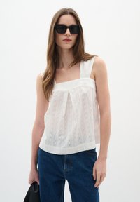 Haut sans manches blanc avec un col carré, un design plissé et des rayures texturées ; associé à un jean bleu taille haute et des lunettes de soleil noires.