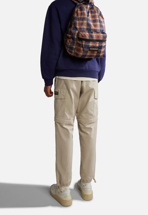 Vue arrière d'une personne portant un sweat-shirt violet foncé, un pantalon cargo beige avec des poches, et des baskets blanches, portant un sac à dos à carreaux.