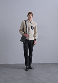 Jachetă dintr-un amestec de lână bej cu accente negre și logo-ul 'EA'. Pantaloni negri și o geantă gri tip crossbody. Căciulă tricotată maro și pantofi negri.