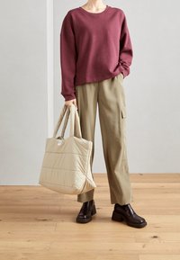 Bordeauxrotes Sweatshirt, beigt gesteppt tote Tasche, olivgrüne weit geschnittene Hose, schwarze klobige Schuhe mit Absatz, Holzboden im Hintergrund.