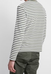 Langärmliges Hemd in olivgrün mit weißen Streifen, strukturierter Stoff, runder Ausschnitt, taillierte Form. Kombiniert mit grünen Hosen mit Gesäßtaschen.