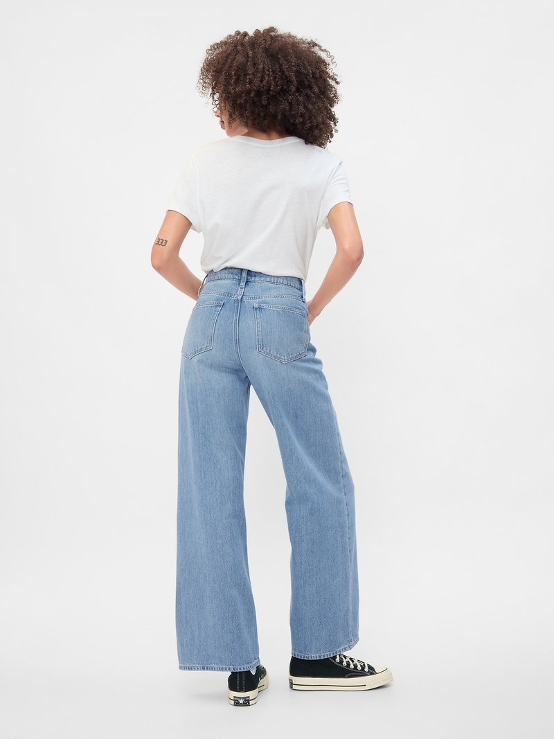 Person mit lockigem Haar steht mit dem Rücken zur Kamera, trägt ein weißes T-Shirt, hellblaue weite Jeans und schwarze High-Top-Sneaker.