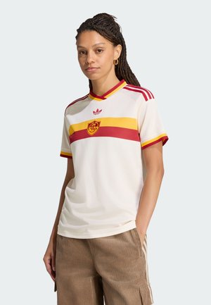 Kvinde med fletninger iført en hvid sportstrøje med røde og gule striber og emblem, kombineret med brune kordfløjlsbukser, står med hånden i lommen.