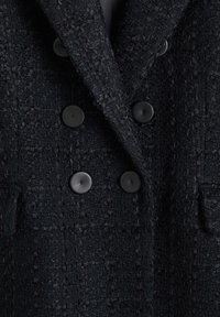 Manteau noir à double boutonnage en tissu texturé, doté de boutons noirs proéminents et de poches plaquées, présentant une silhouette structurée.
