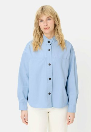 Femme blonde portant une chemise bleu clair ample à boutons et un pantalon blanc, debout devant un fond blanc.