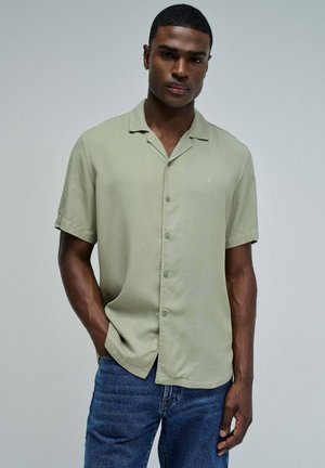 Joven con una camisa verde claro de manga corta y botones y vaqueros azules, de pie con una mano en el bolsillo sobre un fondo liso.