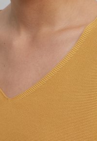 Pullover a V giallo sfatto a maglia con un motivo testurizzato. La vista ravvicinata mette in risalto il collo e i dettagli del tessuto.