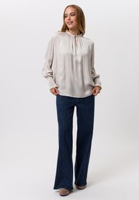 Hellbeige Bluse mit Schlüssellochausschnitt und gerafften Ärmeln, kombiniert mit ausgestellten dunkelblauen Jeans und hellbraunen Schuhen.