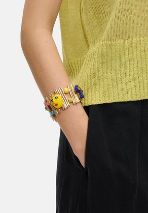 Ręka z kolorową koralikową bransoletką, ubrana w żółty dziergany top i czarne spodnie, dłoń swobodnie umieszczona w kieszeni.