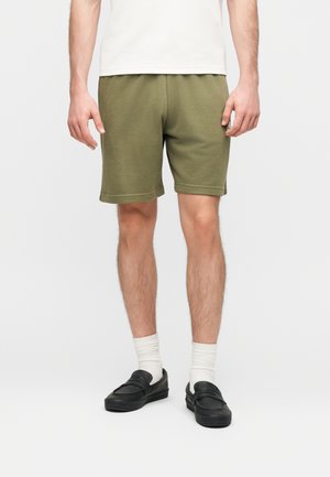 Άνδρας που φοράει χακί σορτς μέχρι το γόνατο, λευκές κάλτσες, μαύρα παπούτσια χωρίς κορδόνια και λευκό πουκάμισο, στέκεται μπροστά σε απλό φόντο.