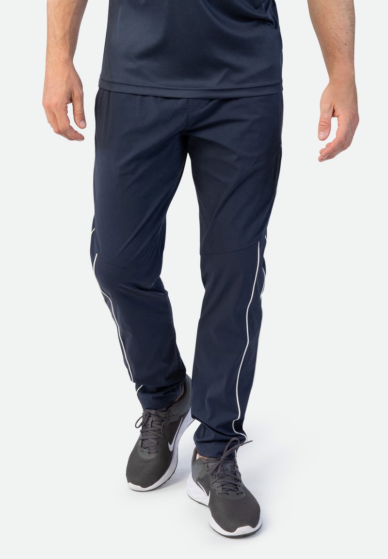 Marineblaue Jogginghose aus leichtem Material mit weißen seitlichen Streifen, elastischem Bund und lockerer Passform. Akzentuiert mit grauen Sneakers.