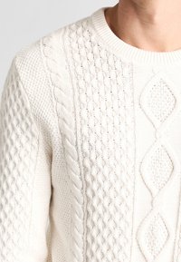 Pull en maille torsadée blanche avec motifs entrelacés et en losange, porté par une personne montrant la zone du cou et des épaules.