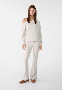 Světle šedá off-shoulder mikina spárovaná s odpovídajícími rozšířenými kalhotami, obojí z měkké látky. Model má na sobě hnědé tenisky. Jednoduchý design.