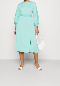 En mintgrön midiklänning med ballongärmar, bälte i midjan och en framspricka, som är kombinerad med vita klacksandaler och en liten handväska.