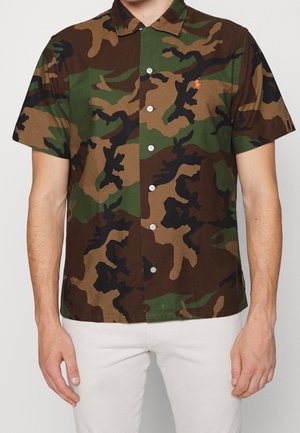 Mann trägt ein kurzärmeliges Hemd mit Knöpfen, das ein Tarnmuster in Grün, Braun und Schwarz sowie ein kleines orangefarbenes Logo auf der Brust zeigt, kombiniert mit hellen Hosen.