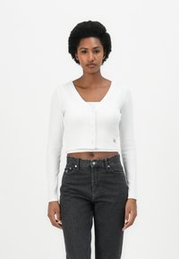 Hvit kortermet cardigan med V-hals og knappelukking, parret med høyt liv mørke jeans. Strikket tekstur med lange ermer.